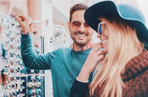 Young man and woman dressed fashionably and wearing sunglasses smiling at each other while browsing a wall of display glasses