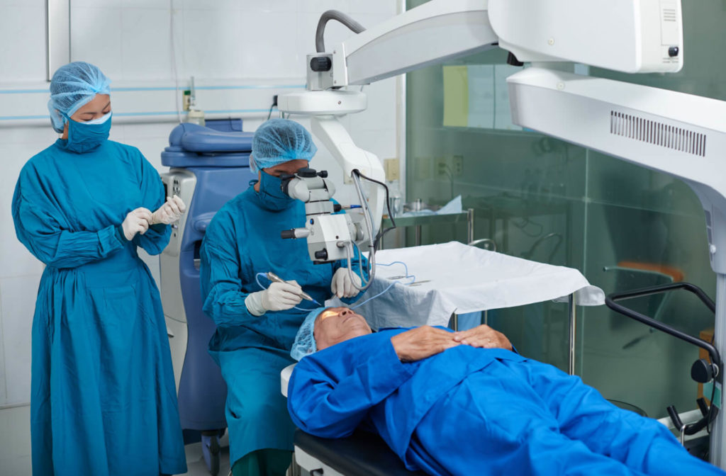 A doctor wearing a blue surgical gown, sitting on a surgical stool beside her female assistant holding a pen-like apparatus in his right hand performing eye surgery on a patient lying face upwards.