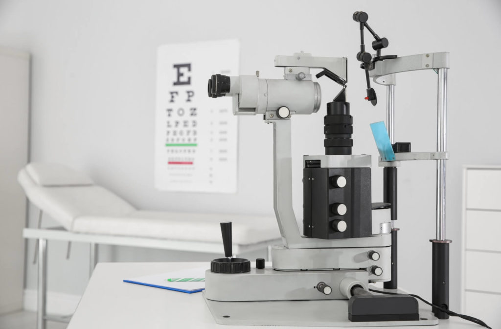 Inside an optometry clinic, a slit lamp on a table and a Snellen chart on the wall.