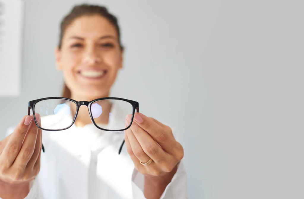 An optometrist holding a pair of prescription glasses as she smiles and looks directly at the camera