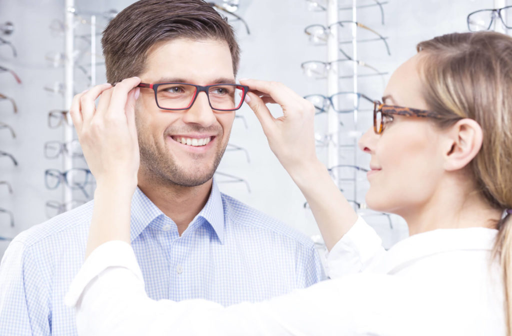 A man smiling and trying on glasses in a store while being assisted by an optician or optometrist.