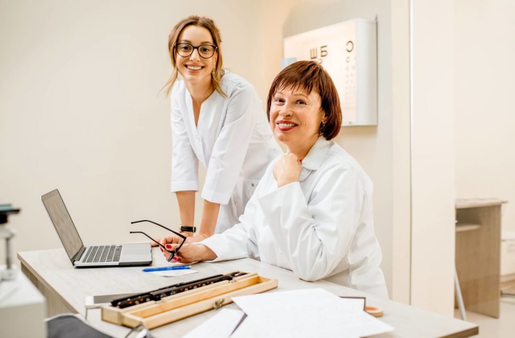 Two female eye care professionals wearing white coats and smiling at the camera while huddled around a laptop.