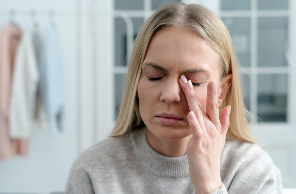 A person with her eyes closed gently presses her fingers to one eye, appearing to experience discomfort or blurriness.